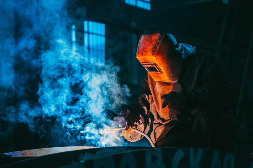 A welder in a workshop using safety gear to weld metal, surrounded by blue smoke and atmospheric lighting.